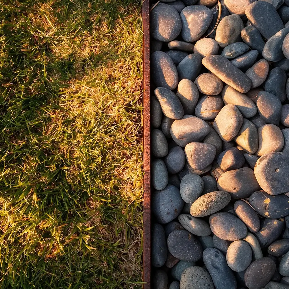Close-up view of a patch of grass on the left and a section of smooth gray stones on the right, divided by a narrow metal border. Warm sunlight casts shadows across both surfaces.
